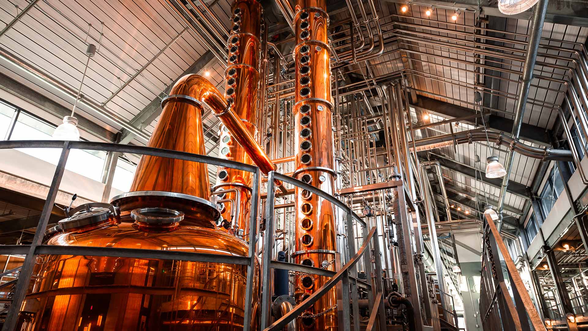 Interior photo of Bently Heritage Distillery|Copper Pot Still|Bently Heritage Bottling Line|Wooden mixing vats|Copper Column Stills inside Bently Heritage Distillery|Entrance to Bently Heritage Distilery|Pot Still|Stainless steel process piping and tanks|Stainless pot and staircase|Green glass bottles lined up on filling line|Bently Heritage Blue Background|||||||||||
