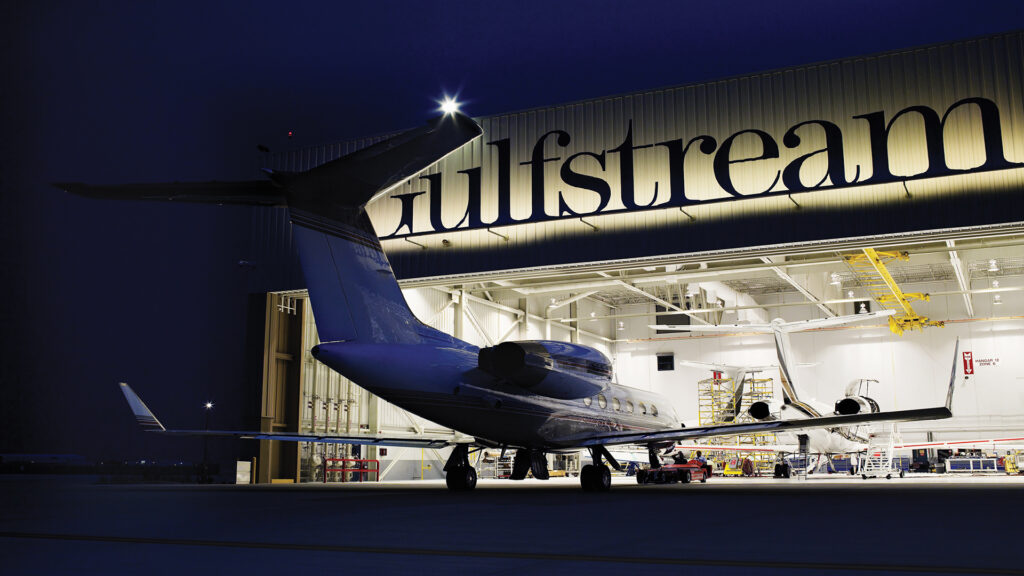Exterior of Gulf Stream Service Center at night. Plane entering hangar.|Exterior of Gulf Stream Service Center at night. Plane entering hangar.|Exterior of Gulfstream Service Center at dusk. Airplane sitting outside of large rectangular building.|Aerial view of Gulfstream Service Center. Rectangular building surrounded by parking lots and parked aircraft.||||