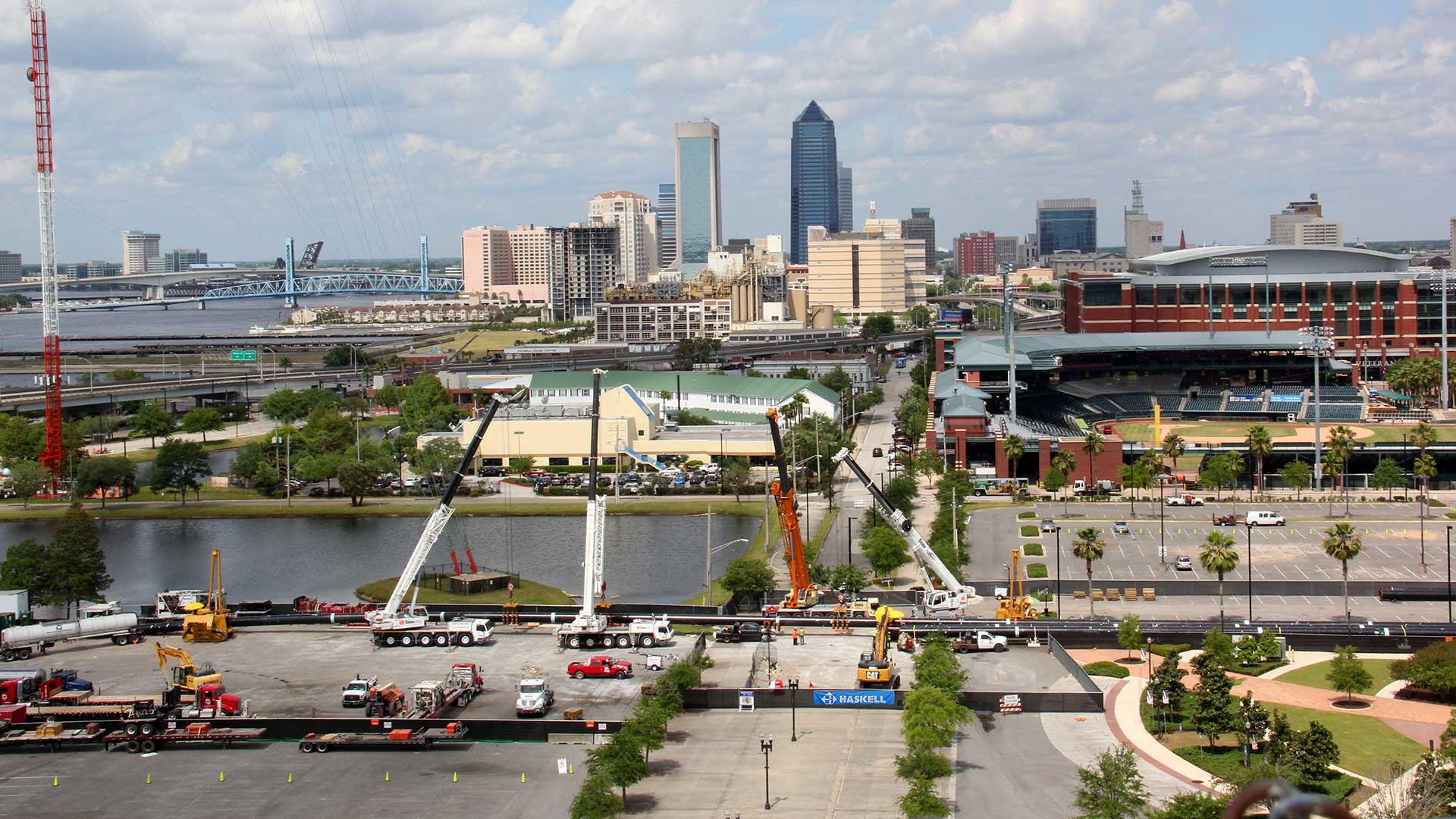 Aerial view of pipe installation in the center of downtown Jacksonville|Giant yellow drill bit|Man inspecting pipe|Pipes on a semi-truck bed|Very long pipe on stands along a highway full of cars. Large tractors
