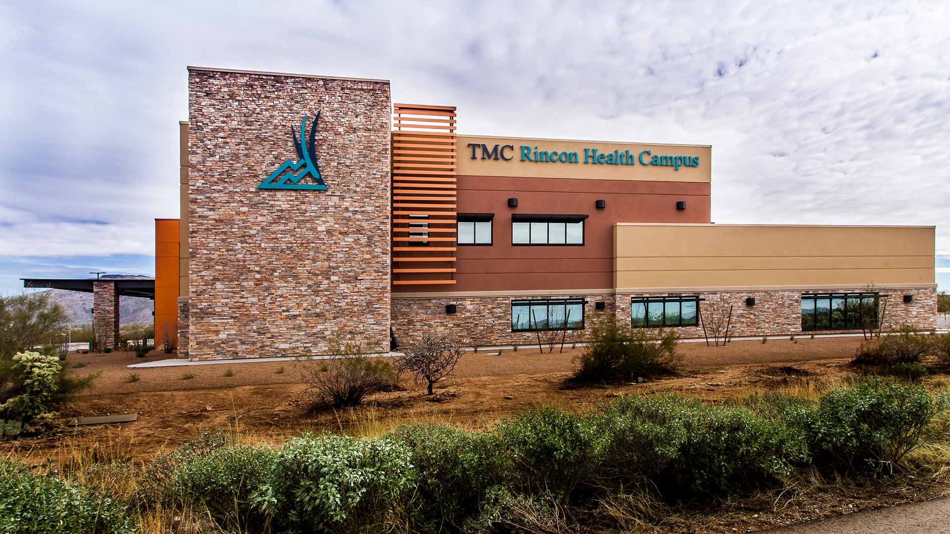 Exterior photo of TMC Rincon Health Campus in the desert. Natural earth tones were used on the exterior finishes|Exterior photo of TMC Rincon Health Campus in the desert. Cactus and rocks are in the foreground of the photo.|Exterior photo of TMC Rincon Health Campus. Street and parking area are in the foreground of the photo.|Exterior photo of TMC Rincon Health Campus. Street is in the foreground of the photo.|Waiting area inside the TMC Rincon Health Campus. Orange chairs and brown tables sit in the middle of the room.|Nurses station and hallway of the TMC Rincon Health Campus|Reception desk of the TMC Rincon Health Campus. Rock wall with large sign with logo are behind the desk.|||||||